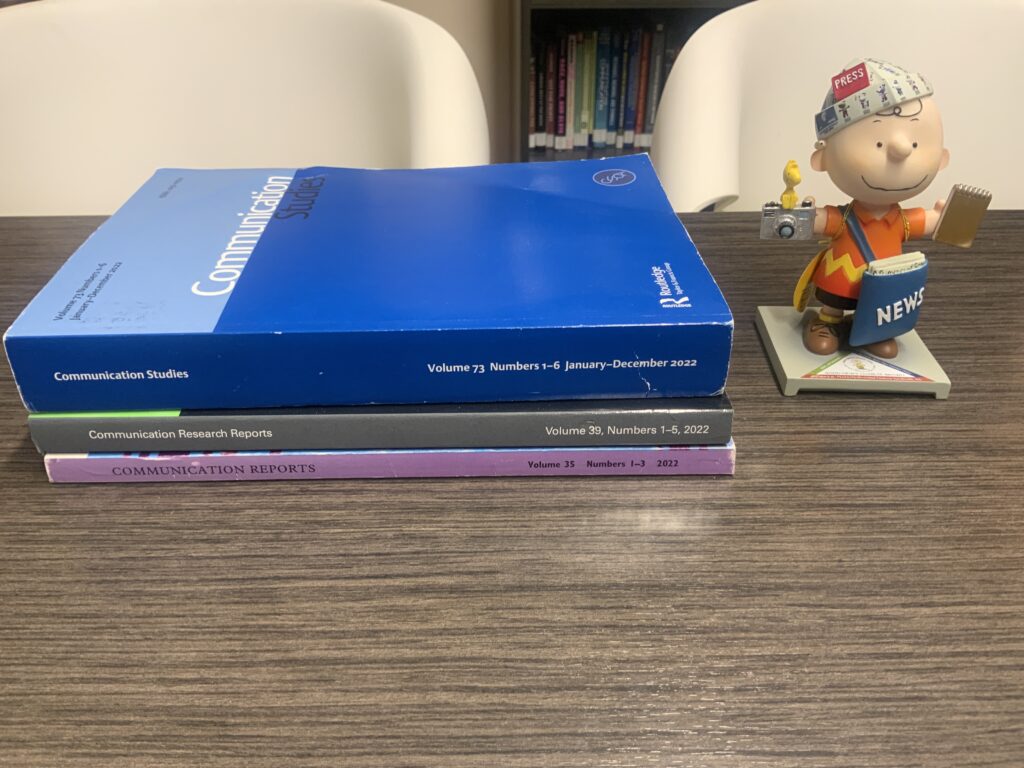 Three journals: Communication Studies, Communication Research Reports, and Communication Reports, along with a statue of Charlie Brown carrying a bag of newspapers, a reporter’s notebook, and a camera with Woodstock on the camera.